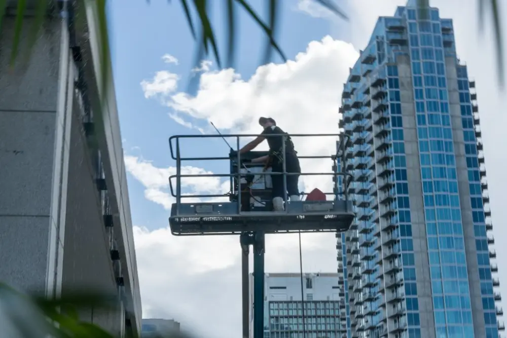 A worker on an aerial lift using commercial pressure washing equipment to clean a high-rise building exterior.